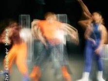 Drei Tänzer*innen bewegen sich dynamisch in verschwommenen, farbenfrohen Kostümen auf einer Bühne.