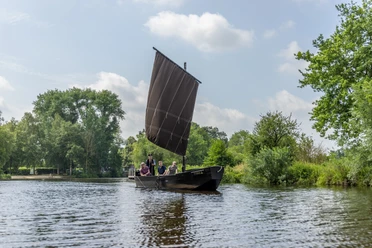 Mit dem Torfkahn auf Hamme Ein traditioneller Torfkahn mit vier Personen segelt auf der Hamme, umgeben von grüner Flusslandschaft.