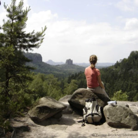 blick-von-affensteinen-c-Sae2-300x300.jpg Eine Person sitzt auf einem Fels und blickt über bewaldete Hügel und Felsen.