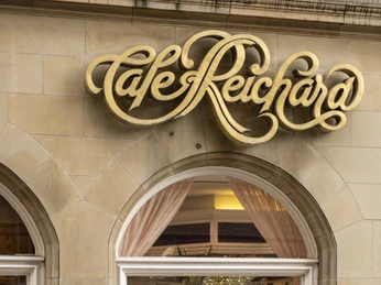 Cafe Reichard Außenansicht mit Blick durch das Schaufenster auf die Tortenvitrine. Darüber in golden geschwungener Schrift Cafe ReichardExterior view with a view of the cake display case through the shop window. Above it, in curved gold lettering, Cafe Reichard