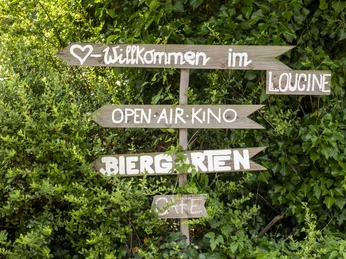 Cinenova Ein hölzernes Wegweiser-Schild im Grünen begrüßt mit Wegweisern zum Open-Air-Kino, Biergarten und Café.A wooden signpost in the greenery welcomes you with directions to the open-air cinema, beer garden and café.