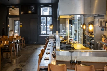 Innenansicht vom Restaurant mit dunklen Wänden und hellen Holzmöbeln. Hinter der Theke, hinter Glas die Küche.