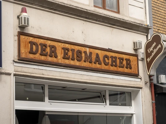 Der Eismacher Fassade eines Geschäfts mit dem Holzschild „Der Eismacher“ über der Eingangstür.Façade of a store with the wooden sign "Der Eismacher" above the entrance door.