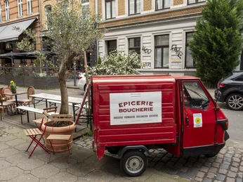Epicerie Boucherie Außengastronomie mit kleinem Olivenbaum. Ein parkendes rotes Piaggio Ape steht präsent im Vordergrund und trägt die Aufschrift vom Cafe.Outdoor dining area with a small olive tree. A parked red Piaggio Ape is prominently in the foreground and has the cafe's logo on it.
