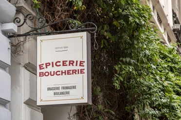 Epicerie Boucherie A sign attached to an ornate metal pole decorates the facade of an old building. Surrounded by green plants.