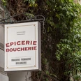 Epicerie Boucherie Ein Schild befestigt an einer verzierten Metallstange schmückt die Fassade eines Altbaus. Umgeben von grünem Gewächs. A sign attached to an ornate metal pole decorates the facade of an old building. Surrounded by green plants.