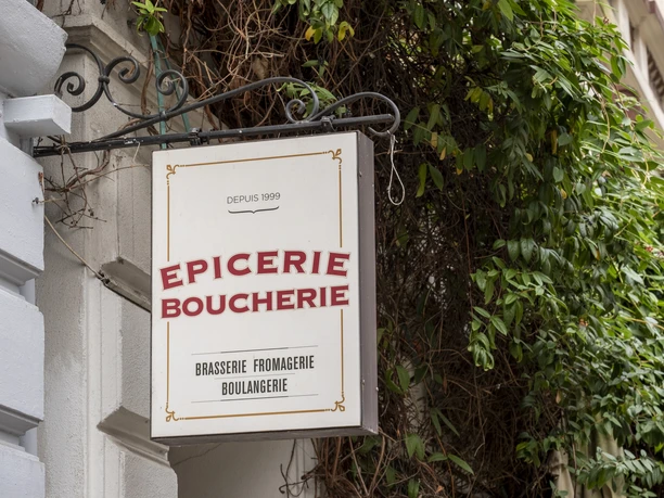 Epicerie Boucherie A sign attached to an ornate metal pole decorates the facade of an old building. Surrounded by green plants.