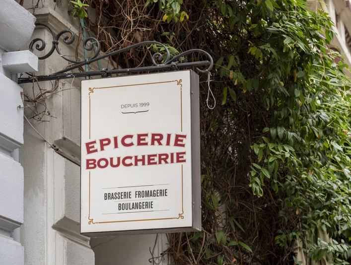 Epicerie Boucherie Ein Schild befestigt an einer verzierten Metallstange schmückt die Fassade eines Altbaus. Umgeben von grünem Gewächs. A sign attached to an ornate metal pole decorates the facade of an old building. Surrounded by green plants.