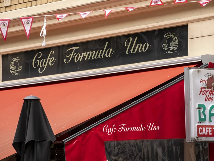 Formula Uno An der Fassade hängt ein schwarzes Schild mit der Aufschrift Formula Uno. Eine Wimpelgirlande flattert im Wind.A black sign with the inscription Formula Uno hangs on the façade. A garland of pennants flutters in the wind.