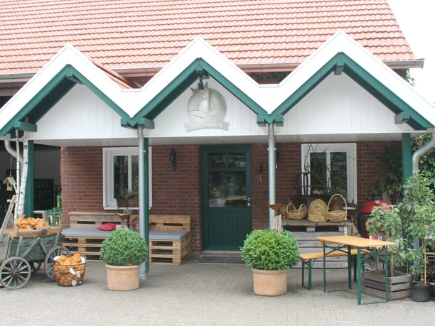 Hof Krone-Raue – Außenansicht Hofladen ©Emsland Tourismus GmbH (4).JPG Eingang des Hofladens mit Pflanzen, Holzbänken und Körben vor rotem Backsteingebäude.