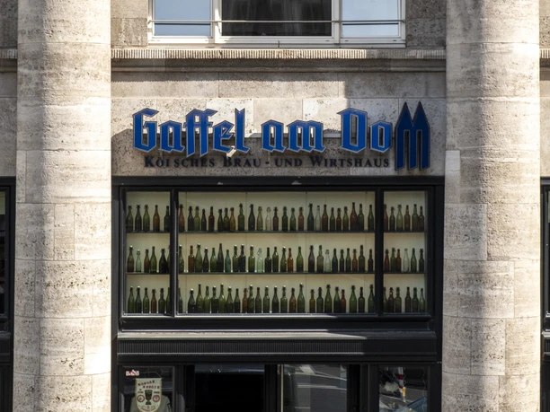 Gaffel am Dom Außenansicht auf das Brauhaus. Zwei markante Säulen rahmen ein Fenster ein, das mit bunten Flaschen dekoriert ist.