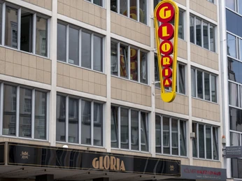 Gloria Das Bild zeigt die Vorderansicht des historischen Gloria Theaters, einem renommierten Veranstaltungsort in Köln. Die Fassade ist durch große Fensterreihen und ein markantes Beleuchtungsschild geprägt, das vertikal an der Gebäudefront angebracht ist. Darunter befindet sich ein Vordach mit dem Schriftzug „GLORIA“ in goldenen Lettern.The image shows the façade of the Gloria Theater in Köln, featuring its prominent red and yellow neon sign against a modern building exterior.