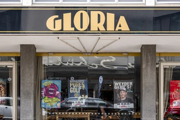 Gloria Schwarzes Schild mit goldener Aufschrift "GLORIA" über einem Kinoeingang mit bunten Filmplakaten.