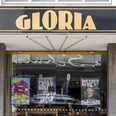 Gloria Schwarzes Schild mit goldener Aufschrift "GLORIA" über einem Kinoeingang mit bunten Filmplakaten.Black sign with gold lettering "GLORIA" above a cinema entrance with colorful movie posters.