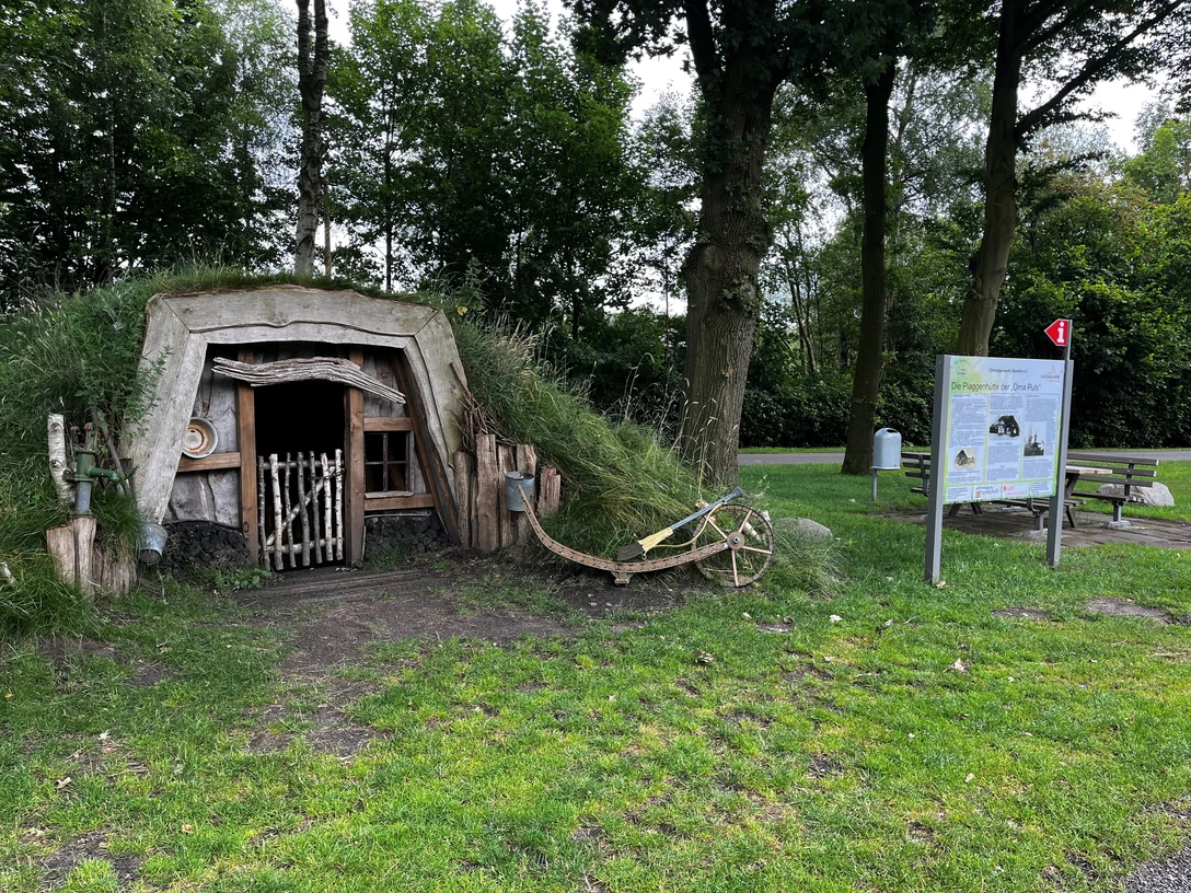 Plaggenhütte Nordloh mit Infotafel.jpg Eine traditionelle Plaggenhütte, neben einem Baum, mit Infotafel und Bank im Grünen.