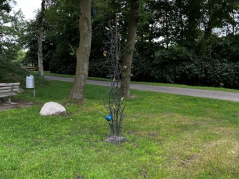 Plaggenhütte Nordloh Lieblingsort 2024.jpg Eine kleine Skulptur aus dünnen Metallstäben steht auf einer Wiese, umrahmt von Bäumen und einem Weg.