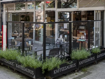 Kaffeesaurus Ansicht auf die Fassade des Café mit Außengastronomie. Ein bestuhlter Bereich ist durch eine transparente Wand vom Gehweg abgetrennt. View of the café façade with outdoor seating. A seating area is separated from the sidewalk by a transparent wall.