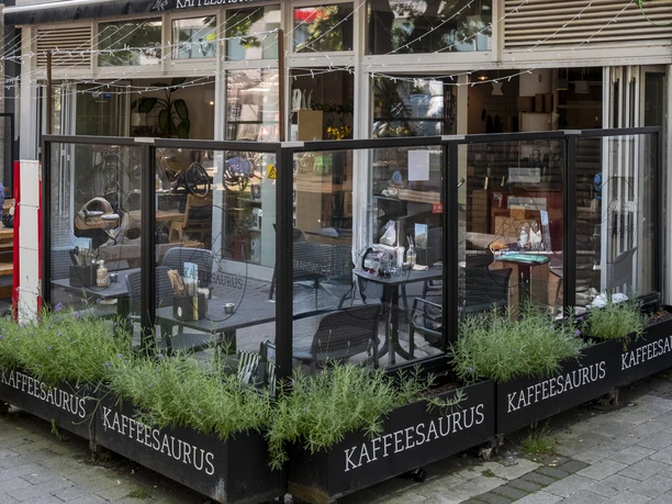Kaffeesaurus View of the café façade with outdoor seating. A seating area is separated from the sidewalk by a transparent wall.