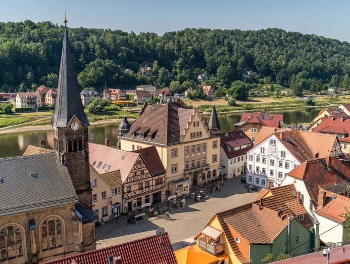 Stadt Wehlen with cyclists' church Historischer Marktplatz mit Fachwerkhäusern und einer Kirche mit Turmuhr, umgeben von grünen Hügeln und einem Fluss bei sonnigem Wetter.Stadt Wehlen with cyclists' churchMěsto Wehlen s kostelem pro cyklistyStad Wehlen met fietserskerk