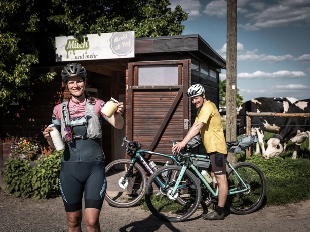 Fresh local products from Milchhof Fiedler Woman in cycling clothes holding milk bottles in front of a farm store, next to her a man with a bicycle; in the background cows in the pasture.
