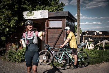 Frische regionale Produkte vom Milchhof Fiedler Frau in Radkleidung hält Milchflaschen vor einem Hofladen, daneben ein Mann mit Fahrrad; im Hintergrund Kühe auf der Weide.