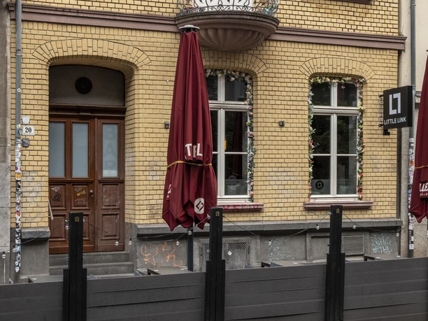 Bar Little Link In front of a bright brick façade stands a red parasol, and a fence made of black-painted wooden planks separates the bar's outdoor dining area from the street.