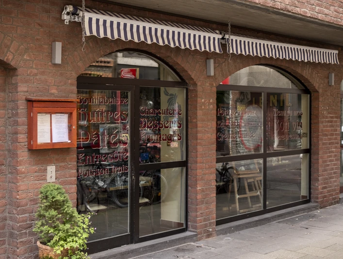 Maison Blue Ansicht auf die Hausfassade mit bodenlanger Fensterfront. Die Fenster sind in verschnörkelter Schrift beschrieben und eine eingefahrene gestreifte Markise befindet sich direkt darüber. View of the house facade with floor-to-ceiling windows. The windows are decorated with ornate lettering and a rolled-up striped awning is located directly above them.