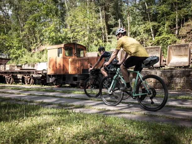 RockHead gravel bikers v muzeu lehké železnice Herrenleite Dva cyklisté s helmami jedou po stezce vedle staré oranžové lokomotivy obklopené zelenými stromy.