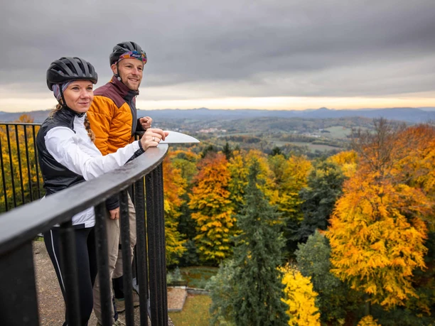 Twee gravelfietsers op de Prinz-Georg-Turm/Ungerberg Twee mensen met fietshelmen staan op een reling en kijken uit over een herfstlandschap met kleurrijke bomen en een bewolkte lucht.