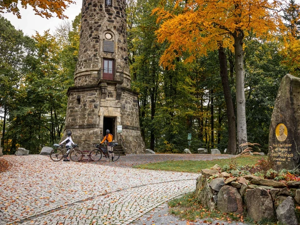 Twee gravelbikers op de Ungerberg in Neustadt i.Sa. Twee fietsers staan voor een historische stenen toren in het herfstbos; kleurrijke bladeren en een gedenksteen zijn zichtbaar.