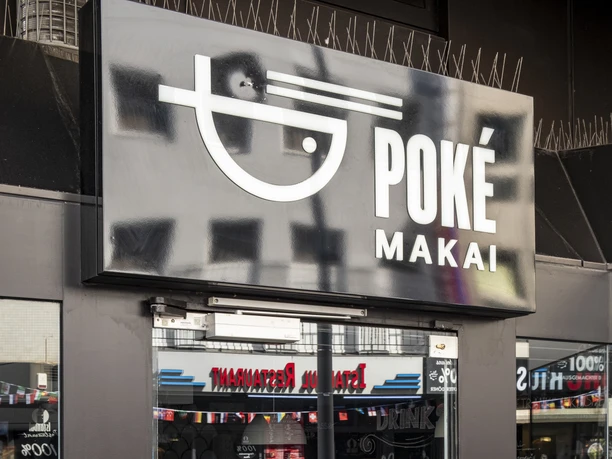 Poke Makai Die Eingangstür von Poké Makai zeigt das Logo des Restaurants und die Öffnungszeiten. Durch die Glasfront sind urbane Straßenszenen sichtbar.