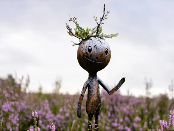 Lächelnde Holzfigur Floki mit Zweigen auf dem Kopf winkt in lila blühender Heide.
