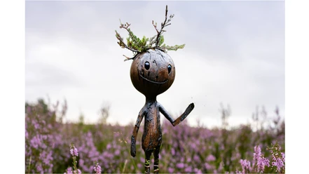 heideaction-mit-floki.jpg Lächelnde Holzfigur Floki mit Zweigen auf dem Kopf winkt in lila blühender Heide.