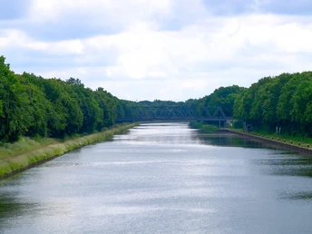 Breiter Kanal von Bäumen gesäumt, ruhiges Wasser, bewölkter Himmel, ferne Brücke am Horizont.