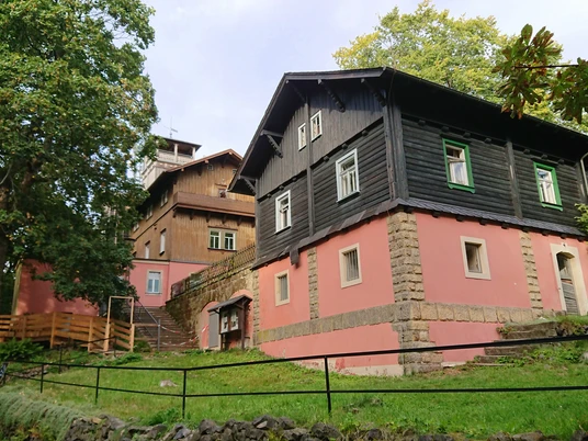 Baude Großer Winterberg Zwei traditionelle Häuser mit rosa und schwarzer Fassade stehen auf einem grünen Hügel, umgeben von Bäumen unter einem bewölkten Himmel.Two traditional houses with pink and black façades stand on a green hill surrounded by trees under a cloudy sky.Dva tradiční domy s růžovou a černou fasádou stojí na zeleném kopci obklopeném stromy pod zamračenou oblohou.Dwa tradycyjne domy z różowymi i czarnymi fasadami stoją na zielonym wzgórzu otoczonym drzewami pod zachmurzonym niebem.Twee traditionele huizen met roze en zwarte gevels staan op een groene heuvel omringd door bomen onder een bewolkte hemel.Due case tradizionali con facciate rosa e nere si ergono su una collina verde circondata da alberi sotto un cielo nuvoloso.