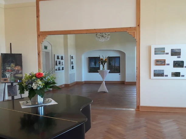 Innenbereich Schloss-Struppen Ein heller Ausstellungsraum mit Holzboden, Gemälden an den Wänden, einem Blumenarrangement auf einem Tisch und einem Kronleuchter an der Decke.