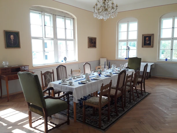Festliche Tafel im Schloss Elegantes Esszimmer mit gedecktem Tisch für acht Personen, umgeben von antiken Stühlen, Kronleuchter und Porträts an den Wänden, helle Atmosphäre.