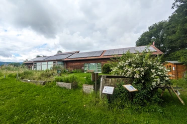 NABU Moorhus Eingebettetes NABU Moorhus im Grünen mit Solarzellen auf dem Dach und umgeben von blühenden Sträuchern.