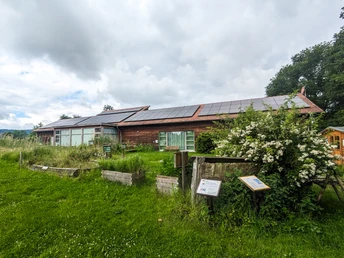 NABU Moorhus Eingebettetes NABU Moorhus im Grünen mit Solarzellen auf dem Dach und umgeben von blühenden Sträuchern.