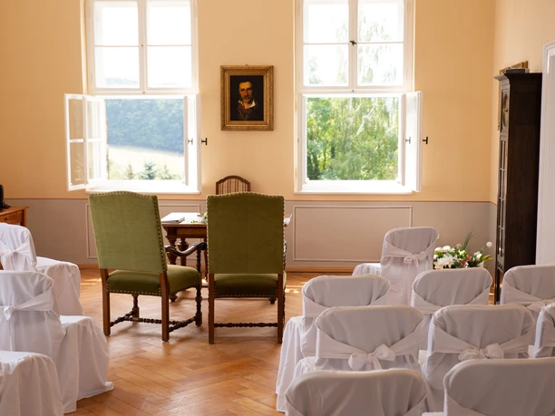 Bright room with two green chairs in front of a table, surrounded by white chairs decorated with bows; window with a view of the greenery.