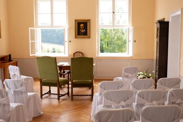 Hochzeitszimmer Schloss Struppen Heller Raum mit zwei grünen Stühlen vor einem Tisch, umgeben von weißen, mit Schleifen dekorierten Stühlen; Fenster mit Blick ins Grüne.
