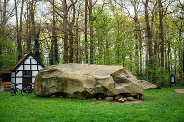 Naturdenkmal Großer Stein in Tonnenheide vor Fachwerkhaus, umgeben von dichten, grünen Bäumen.
