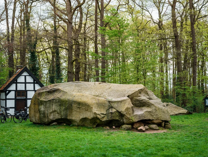Naturdenkmal Großer Stein in Tonnenheide vor Fachwerkhaus, umgeben von dichten, grünen Bäumen.