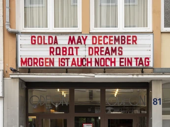 Odeon Das Bild zeigt den Eingang des Odeon Kinos mit einem Vorbau aus Glas. Über der Tür hängen Filmankündigungen in großen roten Buchstaben auf einer weißen Anzeigetafel. Ein Zugangsweg mit sanfter Rampe im Vordergrund verstärkt die Barrierefreiheit für alle Besucher*innen.The picture shows the entrance to the Odeon movie theater with a glass porch. Above the door, movie announcements hang in large red letters on a white display board. An access path with a gentle ramp in the foreground reinforces the accessibility for all visitors.