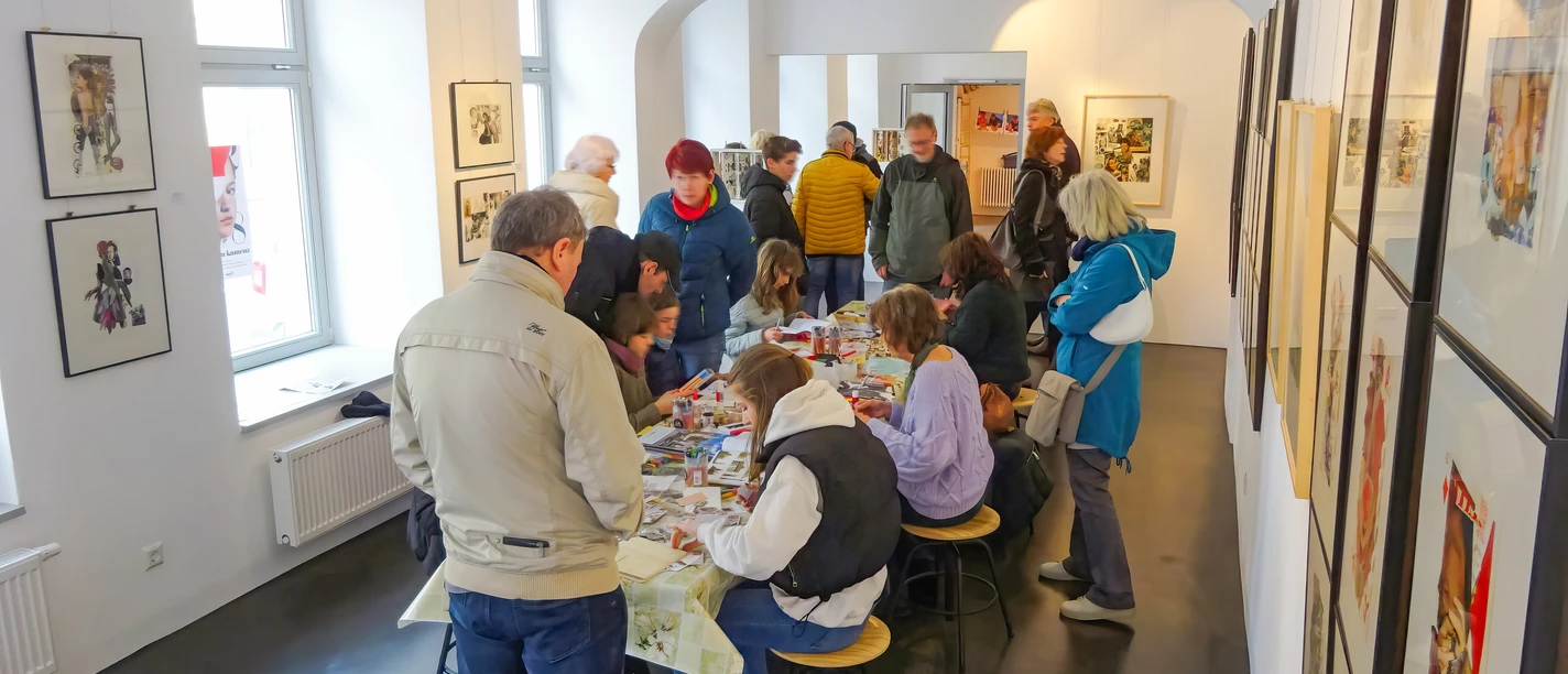 kunstwerksatt-dada-zentrum-kamenz.jpg Menschen in einer Kunstwerkstatt betrachten und gestalten Bilder an einem langen Tisch in einem hellen Raum.