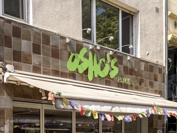 Oshos Place Eine Außenansicht von "Osho's Place" zeigt eine moderne Fassade mit einem Firmenschild in grüner Schrift und bunten Wimpeln. Vor dem Gebäude stehen Tische mit Stühlen, die zum Verweilen einladen. An exterior view of ‘Osho's Place’ shows a modern façade with a company sign in green lettering and colourful pennants. In front of the building are tables and chairs that invite visitors to take a seat.