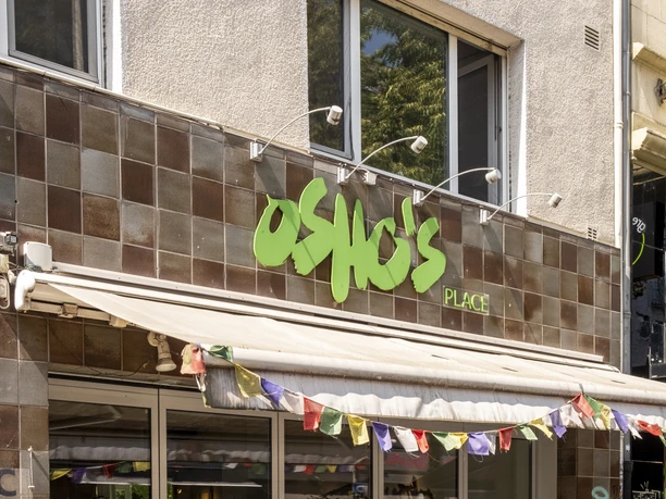 Oshos Place Eine Außenansicht von "Osho's Place" zeigt eine moderne Fassade mit einem Firmenschild in grüner Schrift und bunten Wimpeln. Vor dem Gebäude stehen Tische mit Stühlen, die zum Verweilen einladen.