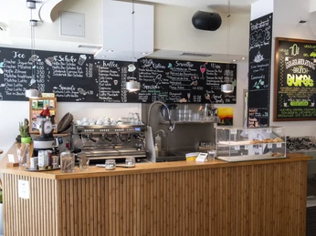 Oshos Place Innenansicht eines Cafés mit einer Bambusverkleideten Theke, darauf stehen eine Kaffeemaschine, Behälter und Tassen. An der Wand sind Kreidetafeln mit handgeschriebenen Menüs zu sehen.Interior view of a café with a bamboo-covered counter, on which there is a coffee machine, containers and cups. Chalkboards with handwritten menus can be seen on the wall.