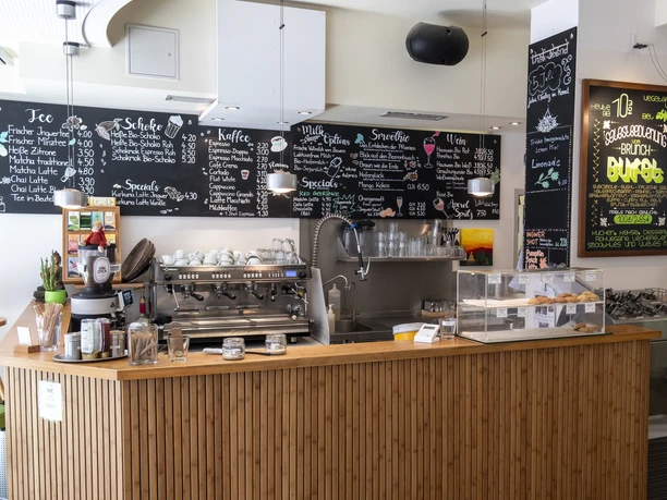 Oshos Place Innenansicht eines Cafés mit einer Bambusverkleideten Theke, darauf stehen eine Kaffeemaschine, Behälter und Tassen. An der Wand sind Kreidetafeln mit handgeschriebenen Menüs zu sehen.