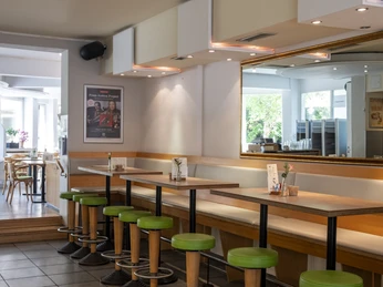 Oshos Place Innenansicht eines modernen Cafés mit hölzernen Stehtischen, grünen Hockern und einer großen Wandspiegelung. Helle Tageslichträume im Hintergrund schaffen eine einladende Atmosphäre.Interior view of a modern café with wooden bar tables, green stools and a large wall mirror. Bright rooms flooded with natural light in the background create an inviting atmosphere.